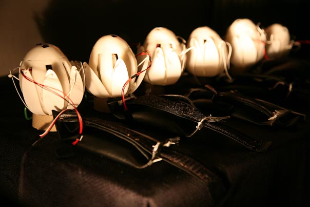 A  photograph of Six  haptic lotus’s in a row from left becoming smaller as they recede to back right. The Lotus’s have a pale ochre tint  against a dark background, and their articulated petals   and string cords are closed,  encircling  an egg shaped  electronic  grilled core.