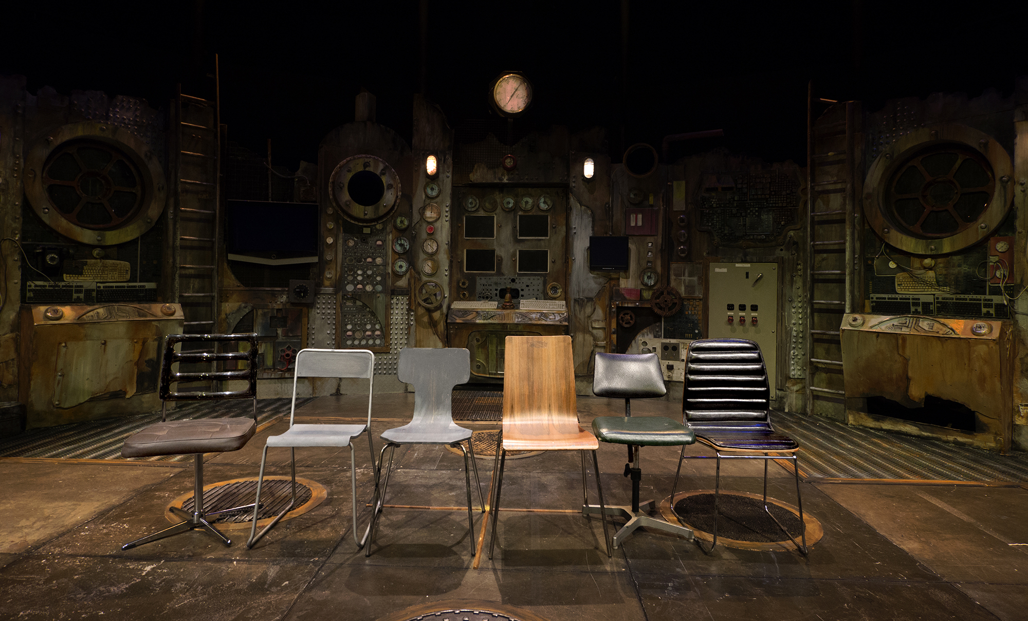   A photograph of 6 upright similar utilitarian empty chairs, Lined up in the foreground centre stage.  Behind is the curved backdrop of dials, controls, consoles, panelled doorways, and port hole windows of the power station.