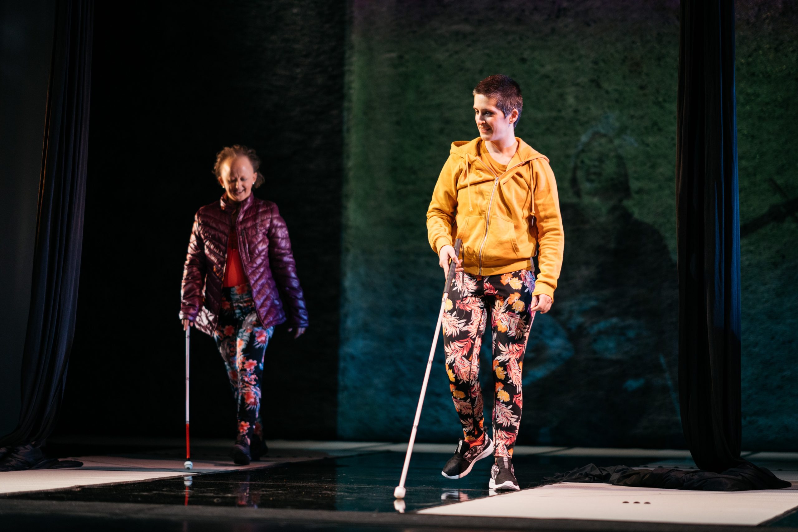 A photograph - Against a dark background, two white performers, Amelia and Sarah, walk forward on stage using white canes. Both are smiling and wearing colourful floral leggings, one with a purple padded outdoor jacket and the other in a yellow hoodie. On the backdrop beyond the open screens behind them, is a waist up blurred image of a Goze. 