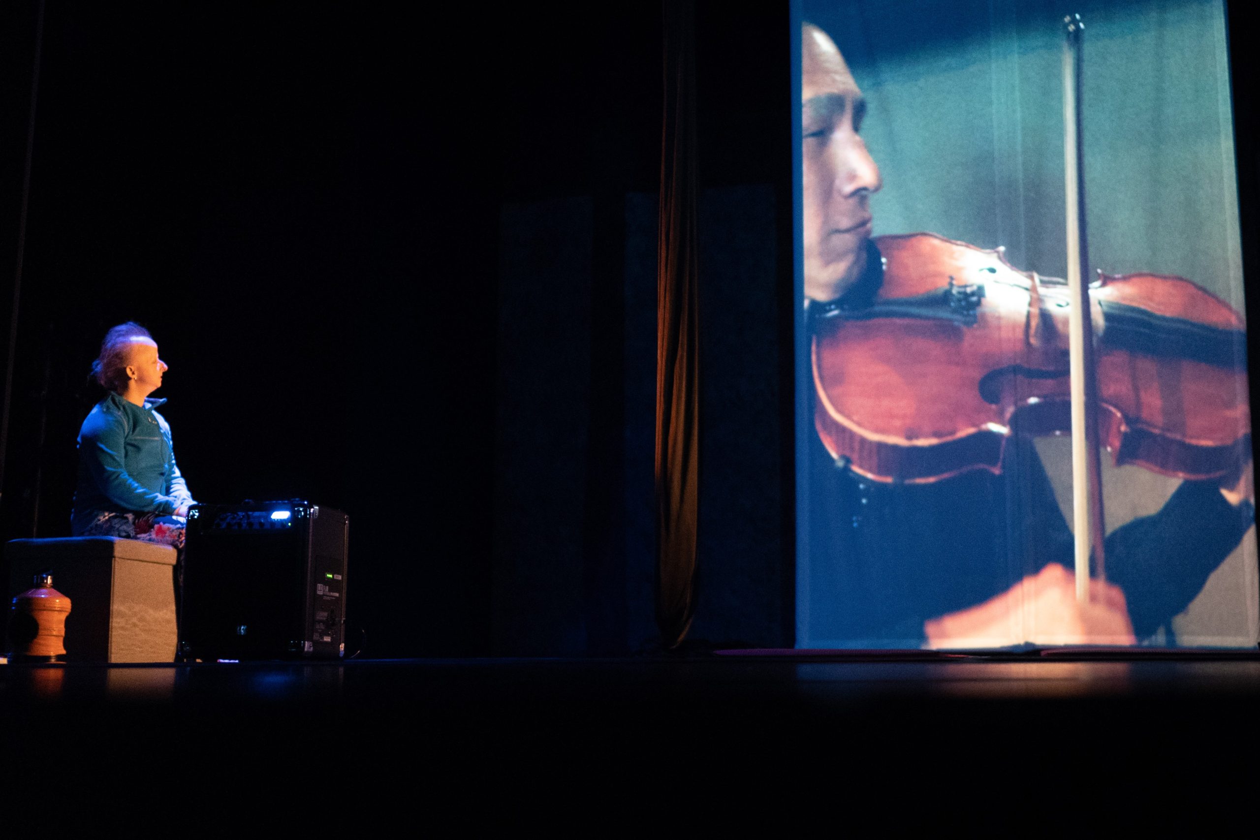 A photograph of a white woman, picked out by a spotlight and seated on a bench   on the far left of the stage, is focused on an East Asian man in profile on a 14ft rectangular screen.  He holds a varnished viola with black fret board visible braced under his chin, and his hand   in the foreground holds a vertical bow.