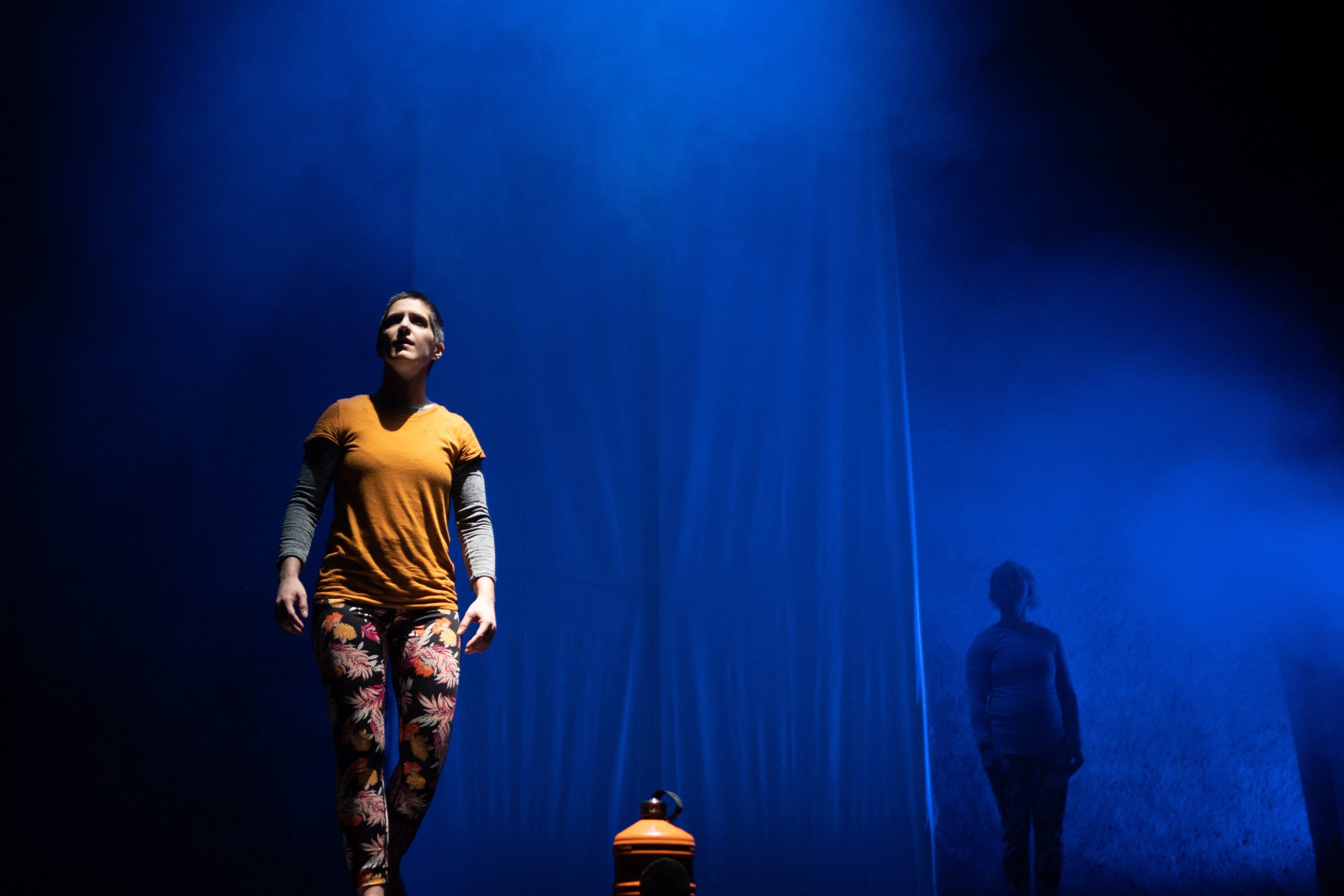 A photograph of Amelia, downstage left, and uplit, only showing from their orange top upwards. Sarah further back stands   featureless in shadow against a misty mid blue light that gradates into darkness at the top and bottom.   In the foreground, centre, is an orange water bottle, clearly lit at the top, but with the bottom disappearing into shadow.