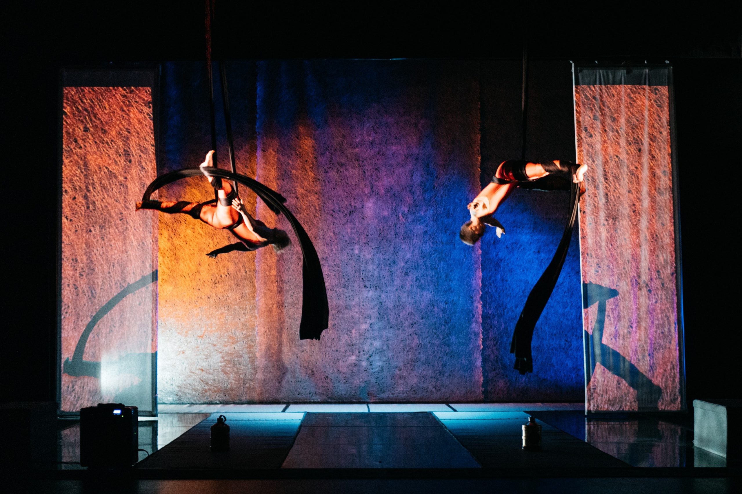 A photograph shows Amelia and Sarah in red skintight body suits, mid fall. Black aerial silks are wrapped around their waists and legs with swathes of the silks sweeping towards the ground. They are both upside down, with heads diagonally pointing towards each other and their legs and arms open in a star shape. To either side of them, on left and right, are long screens with projected coral textured affects, and indistinct shadows of the silks playing on them. Behind the screens, there are three sections of backdrop, with three sections of different colour; On the left orange gradates into darkness at the top, in the middle a Pinky blue gradates into darkness and on the right a vibrant blue gradates upwards into blackness. The black lacquered set floor reflects some of the projections and lighting effects.