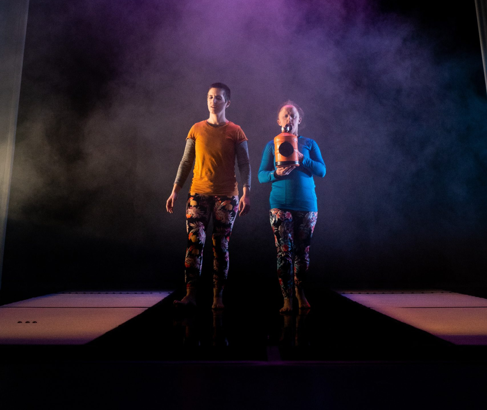  A photograph, taken low angle and front on in grey misty lighting of Amelia on   the left wearing an orange top and floral leggings striding alongside Sarah, centre stage, wearing a blue top and floral leggings.  Both performers have solemn expressions and Sarah cradles the orange water bottle in both hands.  