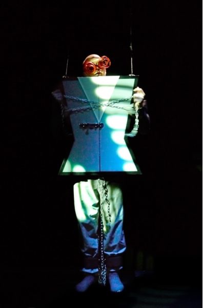 A Caucasian man is standing inside a suspended cabinet box jacket, which is fastened around his body. His head pops through a hole at the top, as do his arms through holes at the side, and he is holding a thick metal chain which is wrapped around the box.