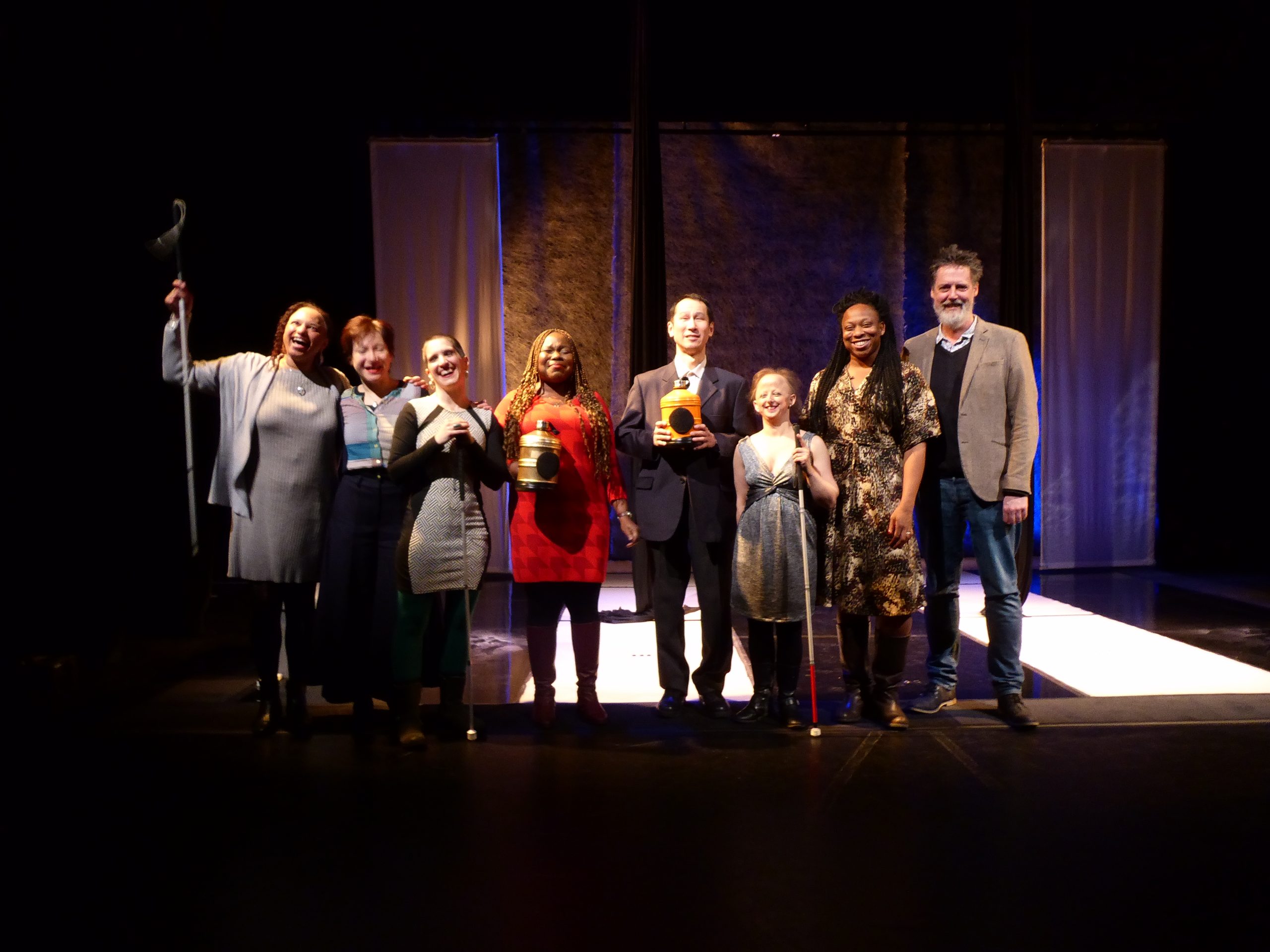 A photograph of the cast and creative team of Flightpaths lined up all smiling and laughing on the stage of the set. The two black silks are behind them and further beyond is the backdrop divided by two screens.  From left to right are Maria in a grey dress, Kumiko in a long dark skirt, Amelia in a panelled black and white checked dress, Victoria with curly long brown hair, holding a golden water bottle in front of her, Takashi in a dark suit holds the orange water bottle in front of his chest, Sarah in a grey and ochre cross over gathered dress holds a white cane. Viki, tall with black braids wears a cream dress with black markings, and bearded Glen is taller in a light grey jacket and jeans.