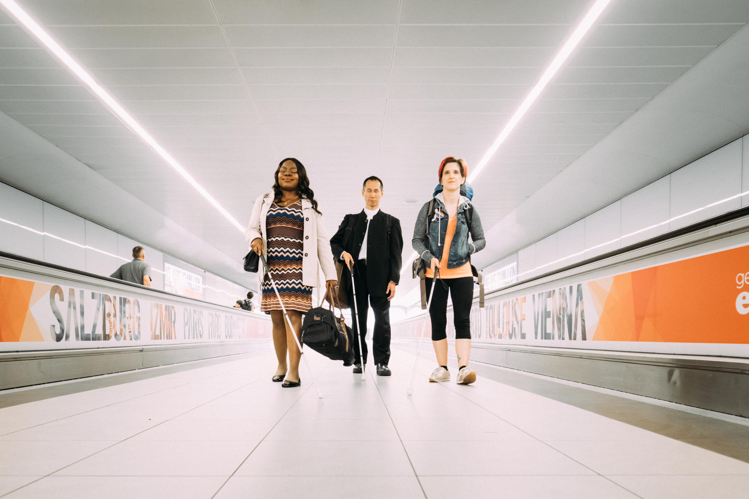  A photograph of Victoria a Nigerian woman, Takashi, a Japanese man and Amelia, an Italian American person, walking towards camera between two airport moving walkways, with their white canes.