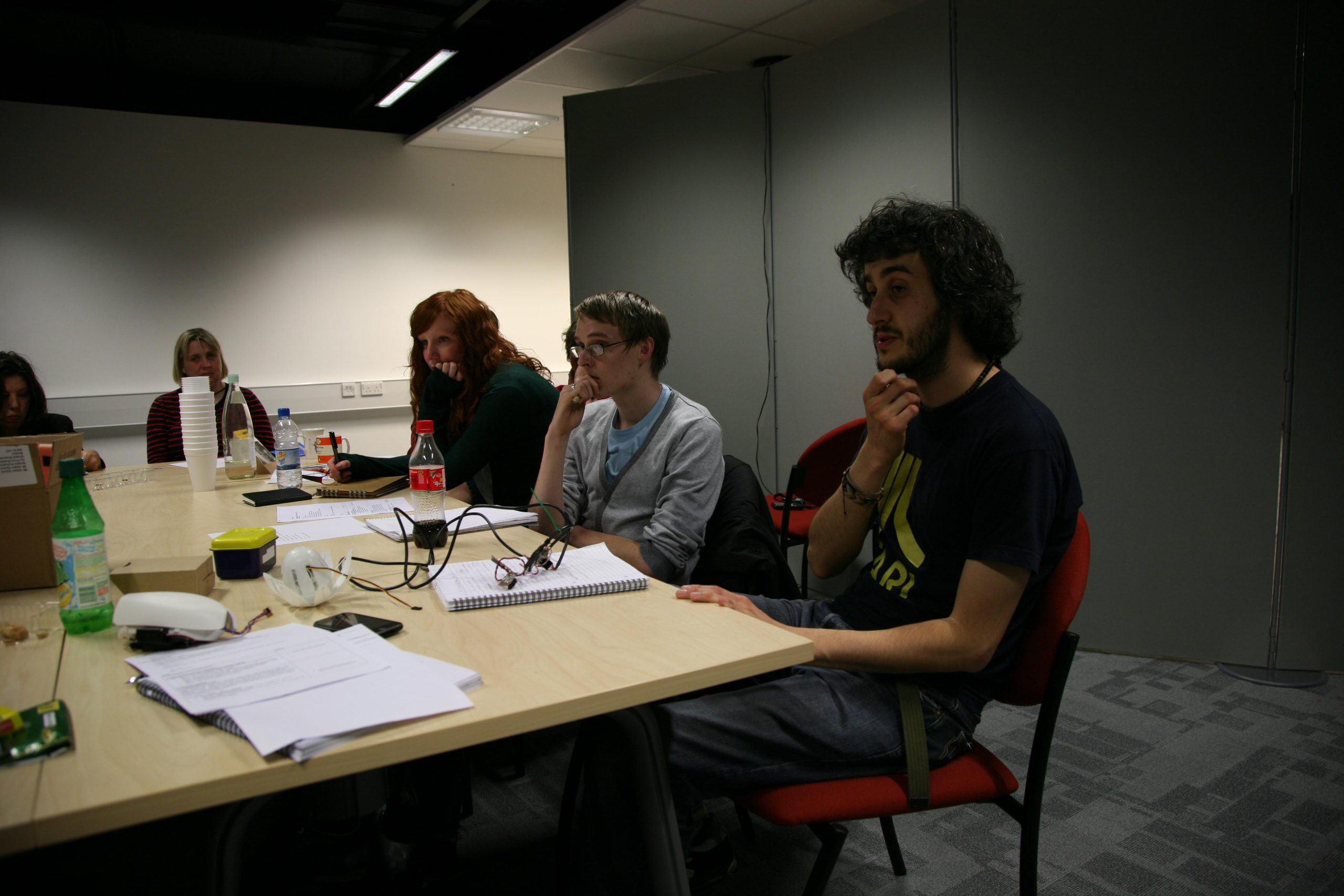 A photograph of several people in discussion at the Open University, seated around a large table, thinking, in discussion, and taking notes, with the lotus, beacons, notebooks and cups in front of them.