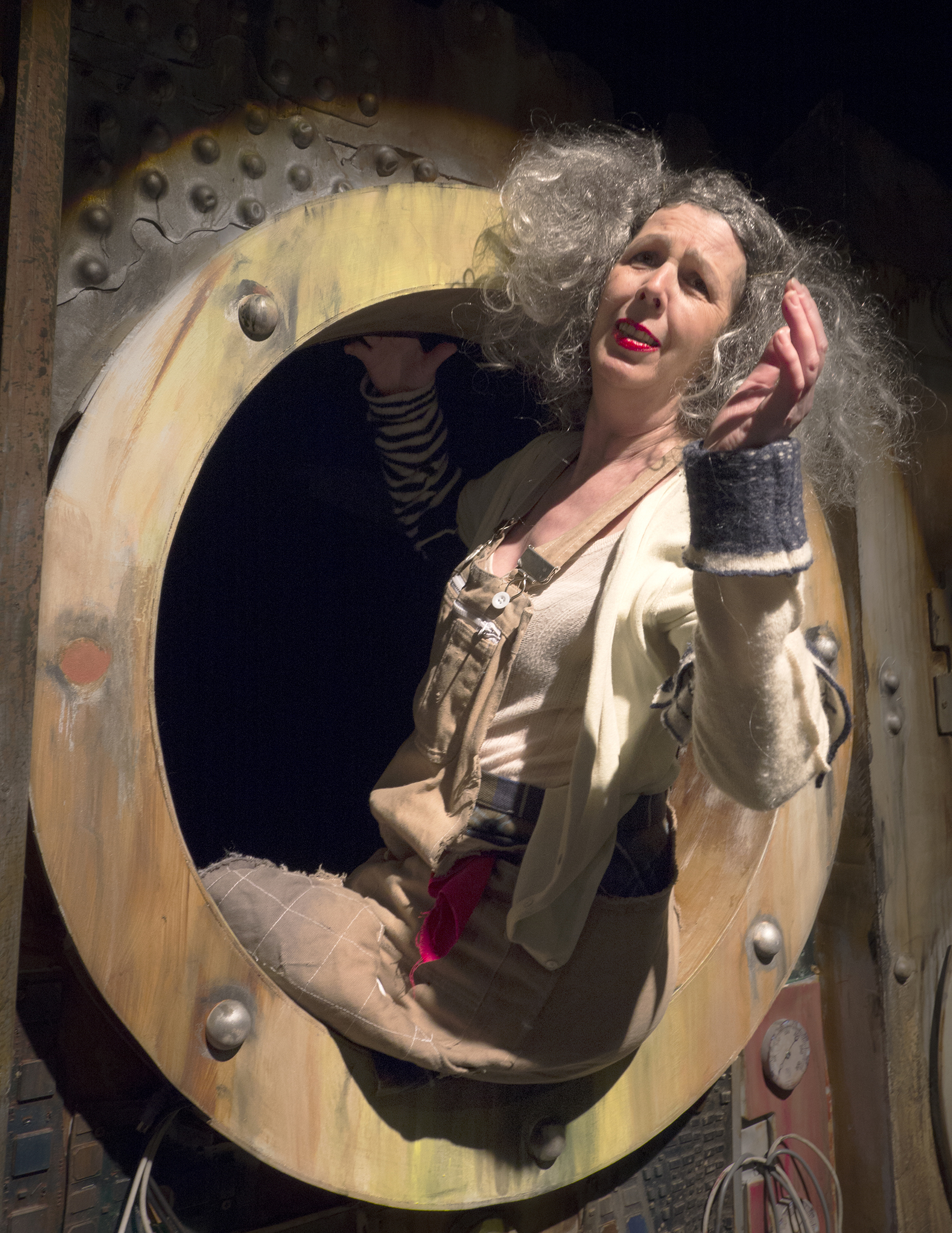 A photograph where one of the porthole windows in the backdrop of the set takes up the whole image. The Old Woman sits on the window cill with both legs hanging outside out of shot, as she turns back to wave. Her voluminous grey hair is teased out on both sides of her head, and she wears bright red lipstick, a brown pinafore dress with patchwork sleeves underneath.