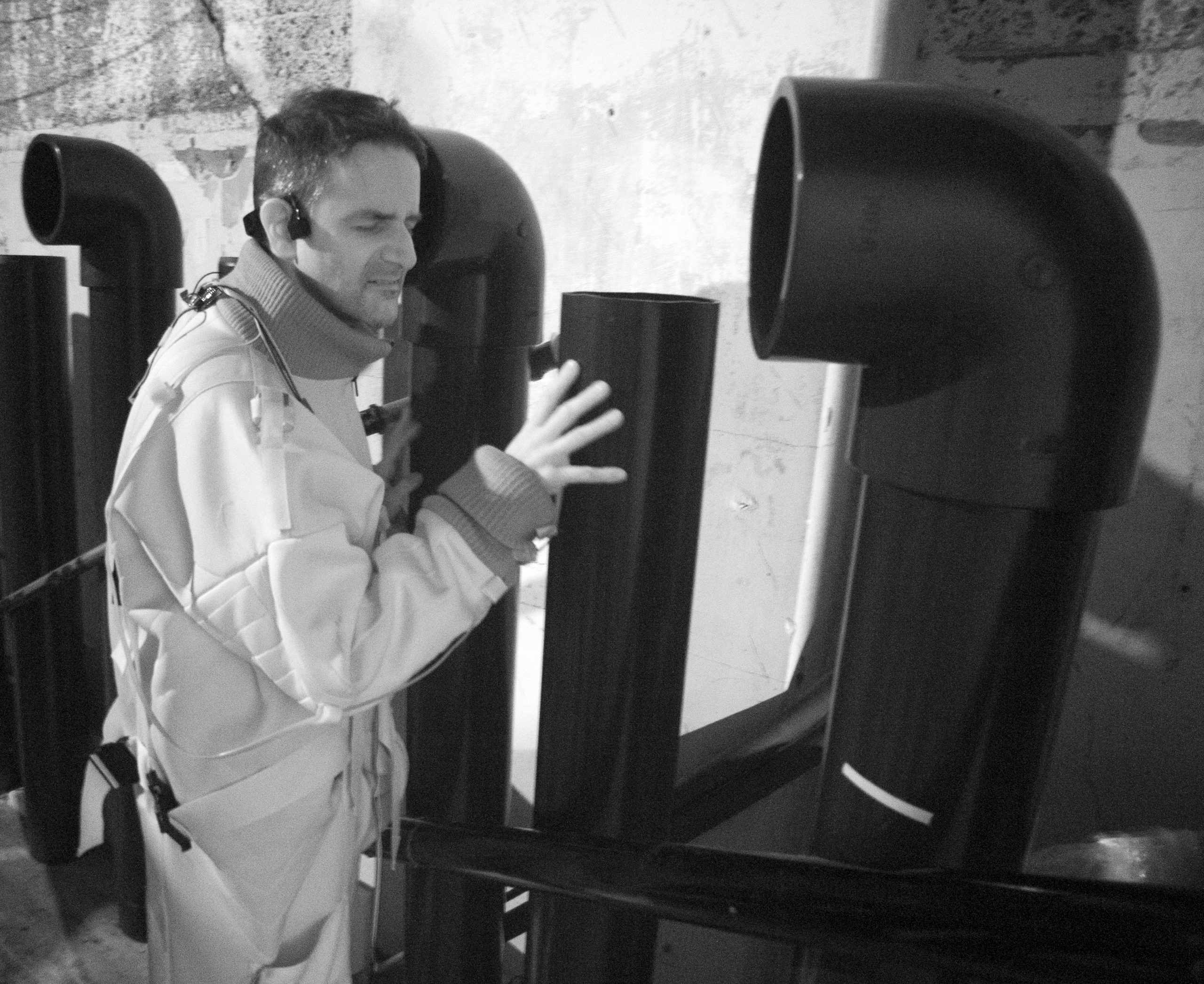 A black and white infra red photograph of the back of a man viewed from over his shoulder. His right hand is touching the wide opening of a foghorn style pipe that is head height. His eyes are closed while wearing bone conducting headphones, as he leans in to listen at the mouth of the pipe.