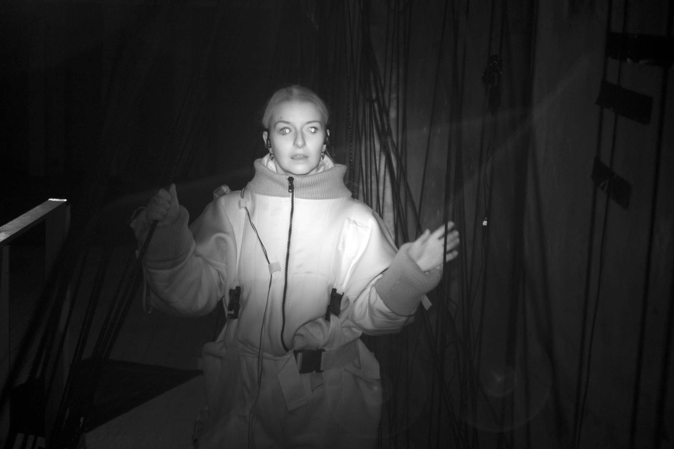 A black and white infra red photograph from front on of a woman surrounded by a tangle of black chords and trailing ropes.  With one hand she touches a rope and grips one with the other, while her eyes are caught by the camera as if alight.
