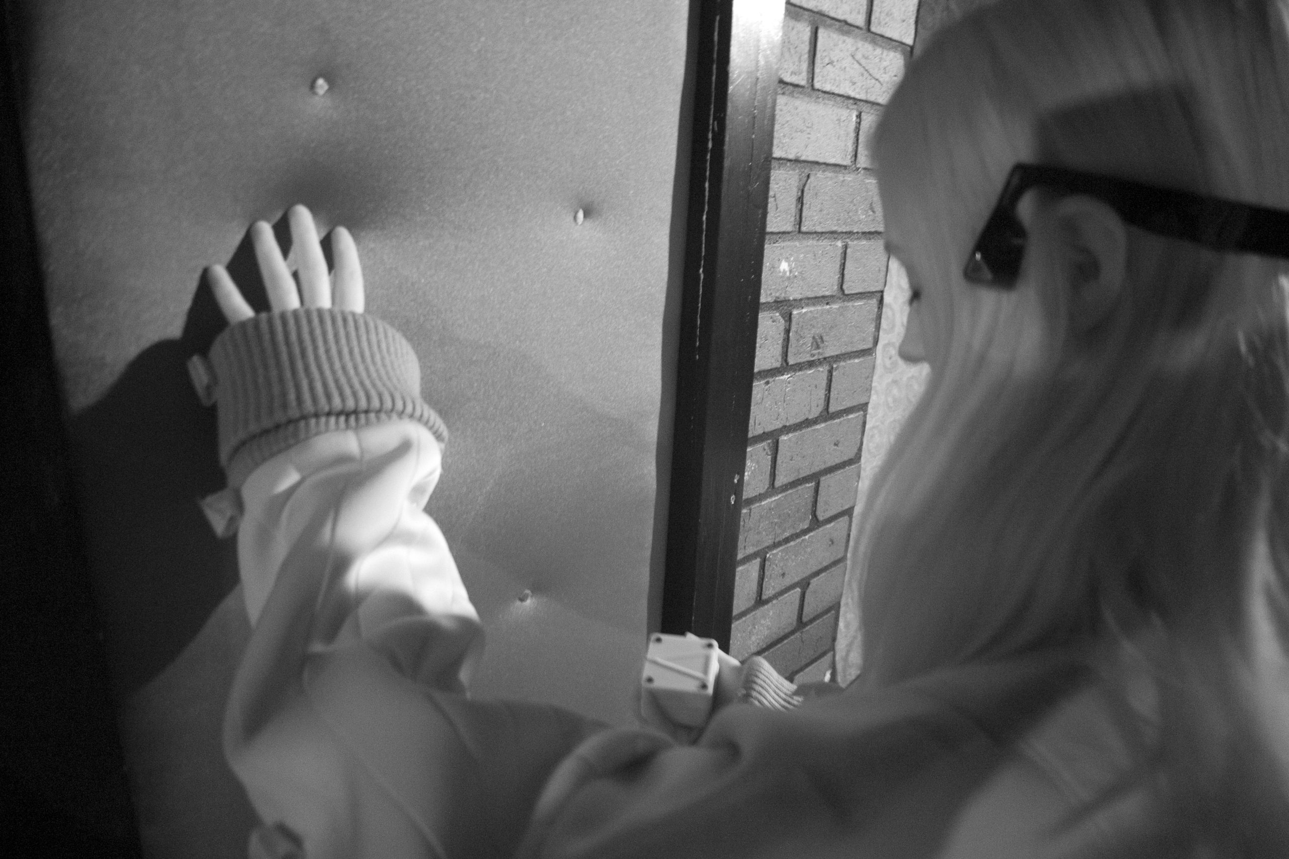 A black and white infra red photograph from over a figure’s shoulder, who’s left arm is extended to feel a window shaped panel in front of them. E-textile dots are sewn into the pane, which vibrate and represent one dimensional flatland women. In their right hand they hold the Animotus that has an arrow on top of it pointing towards the window. 