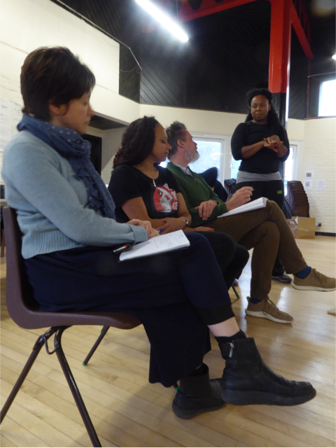 A photograph of Kumiko, a mixed heritage woman, Maria, a mixed heritage woman and Glen, a white man, all in profile on a row of chairs in a vaulted rehearsal venue. All three are focused upwards towards Vicki, a black woman, who stands at the end of the row looking down at them with her hands clasped.