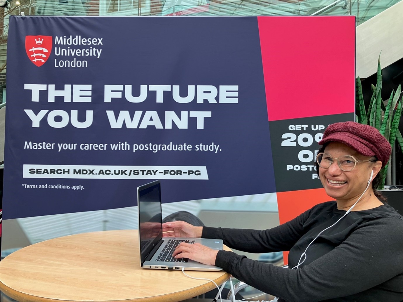 Maria, a biracial middle aged woman, wearing a black long-sleeved top, burgundy peaked cap and glasses, sits at a round table with an open laptop, smiling out at camera, next to a blue banner with the Middlesex University coat of arms which has in capital letters, ' The Future You Want - Master your career with postgraduate study''. Protactile Description: Maria has a bold, playful, talkative energy, sometimes sour, sometimes sweet – She listens, laughs, and moves with definite intention; her long fingered hands give a firm handshake.