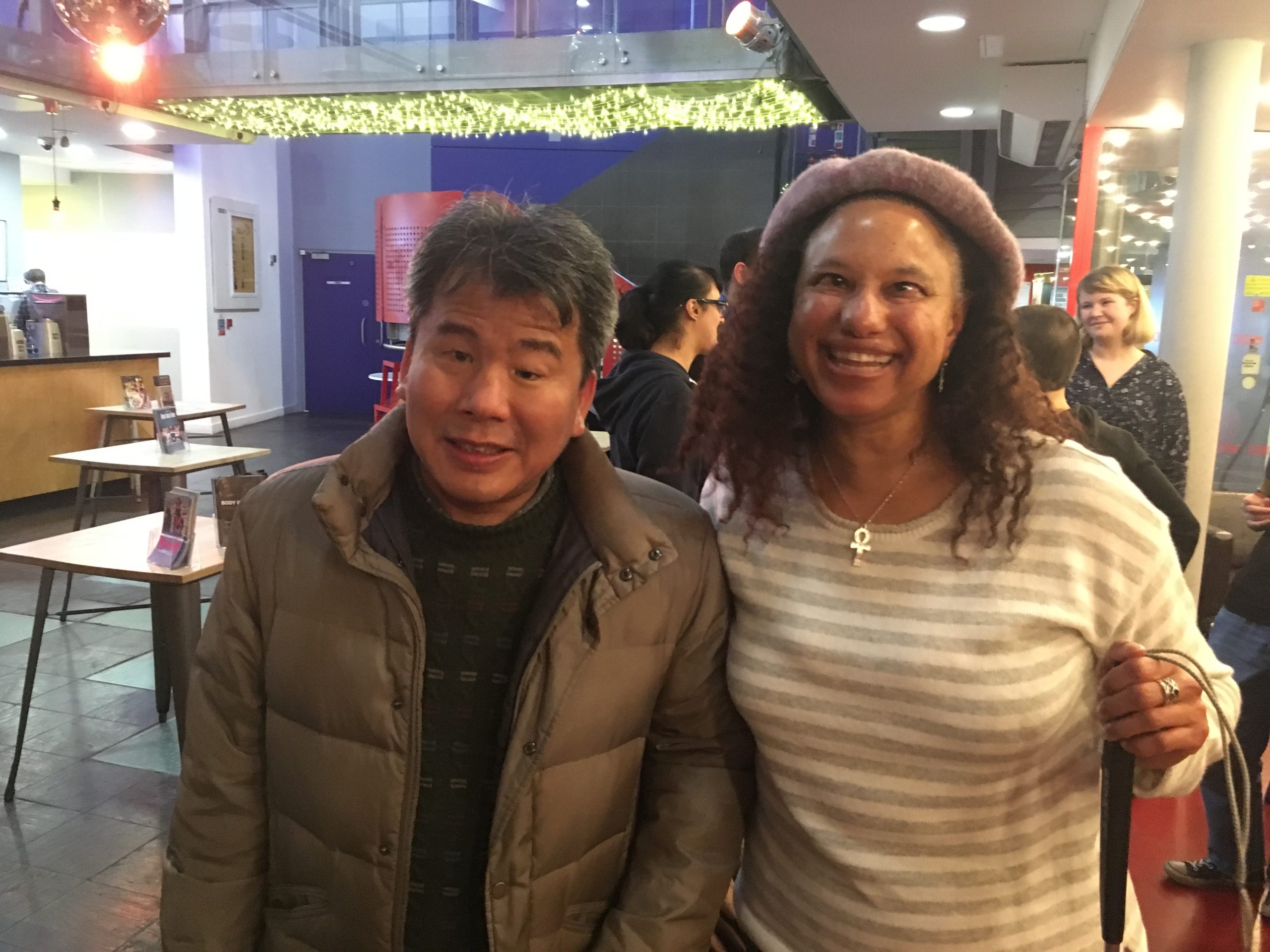 A photograph of Professor Hirose and Maria   both smiling in the café bar of Stratford Circus. On the left Kojiro Hirose, a middle aged Southeast Asian man, wears an olive-green parker jacket with dark green jumper underneath. He has short dark hair with a loose curl sweeping across his forehead.  Next to him on the right, Maria wears a   cream jumper with grey stripes and a mauve beret on top of shoulder length curly brown hair