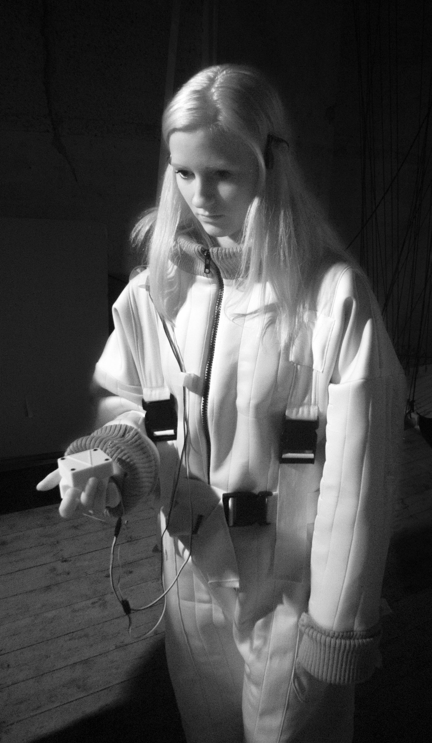 A black and white infra red photograph of a young woman wearing a Flatland quilted and buckled padded uniform, which appears white in this image against a dark background, with some floorboards visible. She holds an Animotus in front of her in her right hand, and the top section of the cube has swivelled. Her  other hand is touching the device, and her expression focuses on being guided by it. 