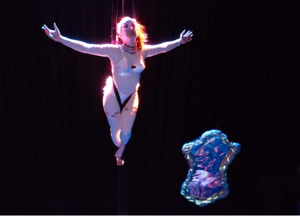 A Caucasian woman is ‘flying’ in mid-air, suspended by her waist. She wears a nude bodysuit and a white pointed bra. She is also wearing an embellished necklace and a half-face mask. Her legs are together and pointed and her arms are flung wide. She is smiling. Beneath her hangs a large glittering fairy tale style mirror.
