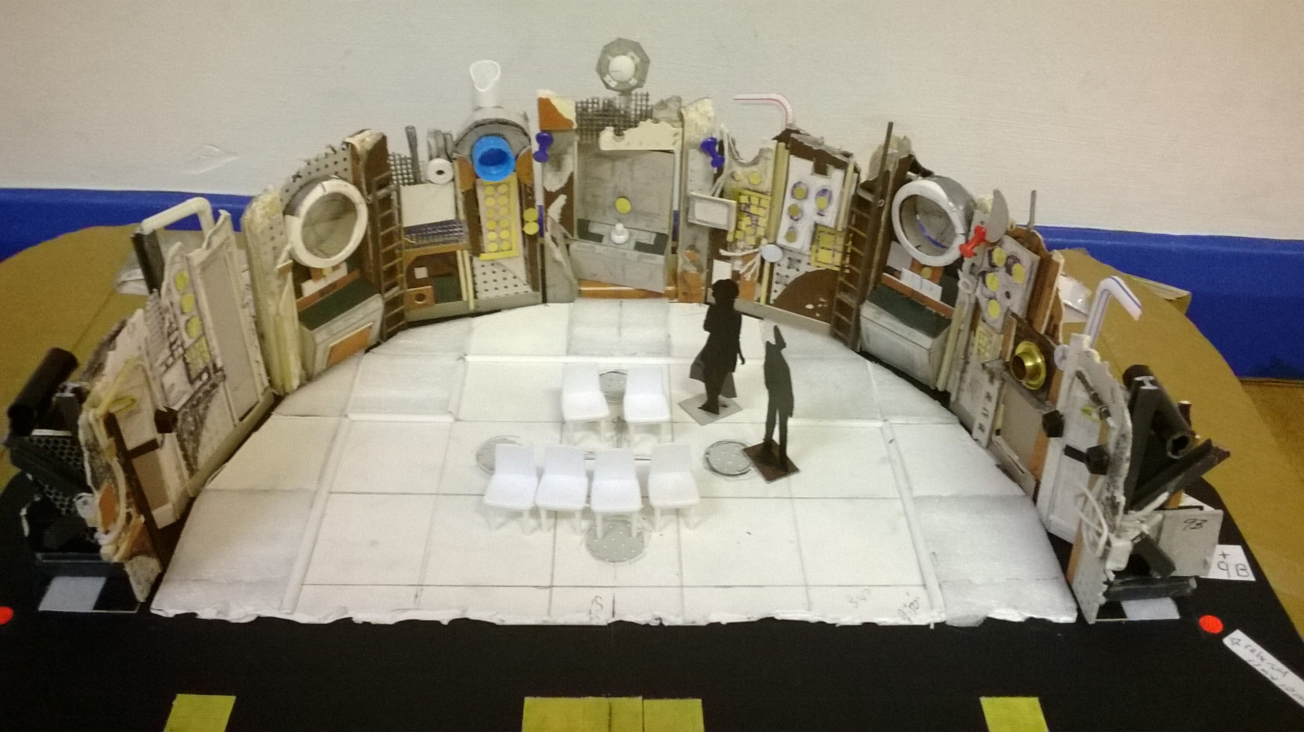 A photograph of the sets model box taken front on and viewed from above. It is a scaled down intricate detailed representation of the set, with a white cardboard semicircle floor marked out in squares, along with a curved wall of the backdrop, made up of panelled doors, ladders, portholes, and controls marked up with a myriad of buttons and make-shift materials. In the centre there are 6 small chairs in a row facing front and two scale black carboard figures to the right of the chairs.