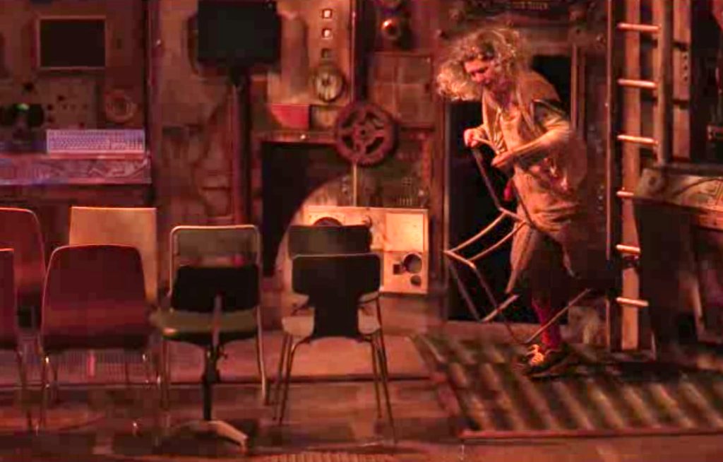 A photograph of two rows of 4 chairs to the left, facing upstage towards the walls of the power station.  On the right, The Old Woman enters with one leg stepped through an open panel in the wall of the power station.  Leaning forward, she carries the empty metal frame of a chair, while all is bathed in a yellowy hue.