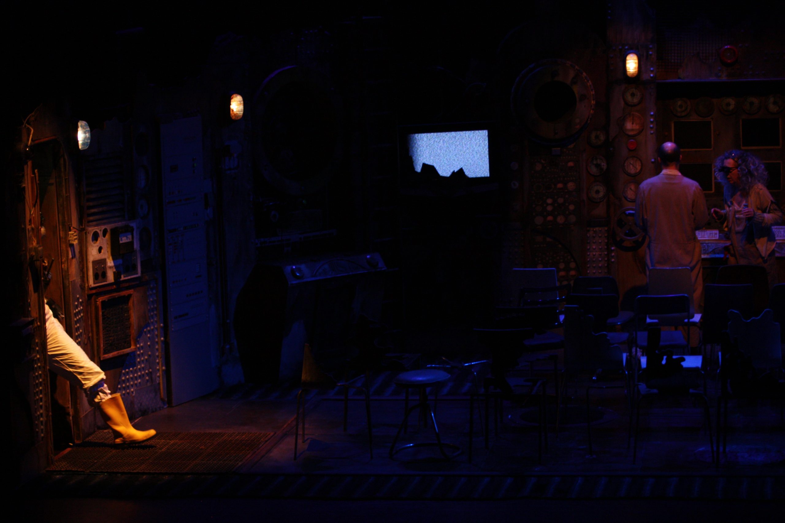A photograph of the dimmed background of the set, with the Old Man and Old Woman in shadow, upstage behind a mass of chairs.  Downstage left is the spot lit white hazmat leg of the Orator, wearing a yellow Wellington boot, as they enter through an open door in the set.