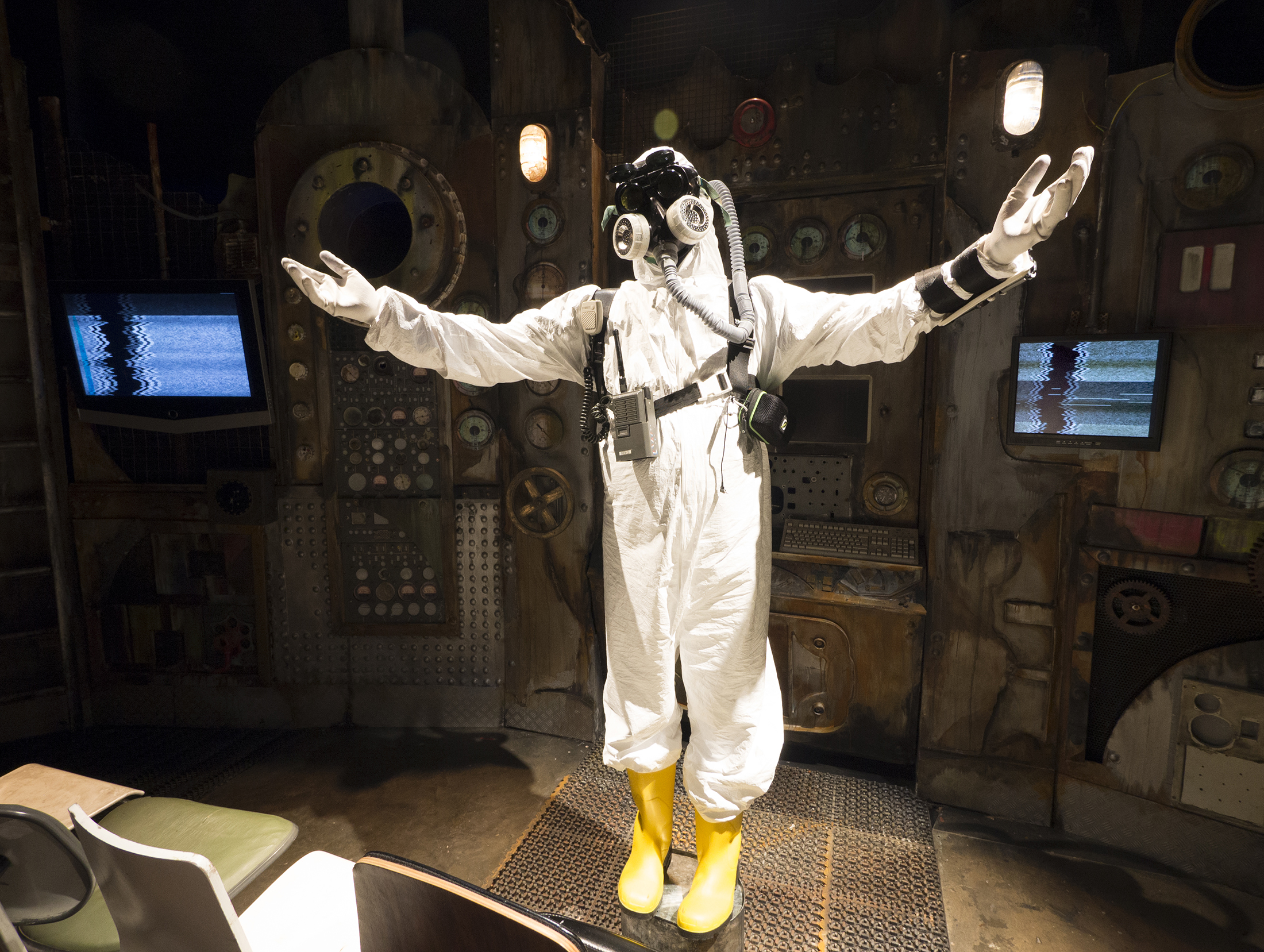 A photograph of a figure Standing on a galvanised bucket with their arms raised out to either side. They are dressed in a white hazmat suit over which a grey harness is strapped, with 3 devices attached to the belt, and a microphone unit fixed to their front. They wear white gloves, yellow boots, along with a full-face mask, with    metal filters connecting to a grey flexible hose that disappears upwards and over their shoulder. Behind them is the backdrop of the power station walls, with two active screens displaying black and white zig-zag lines of interference on them.