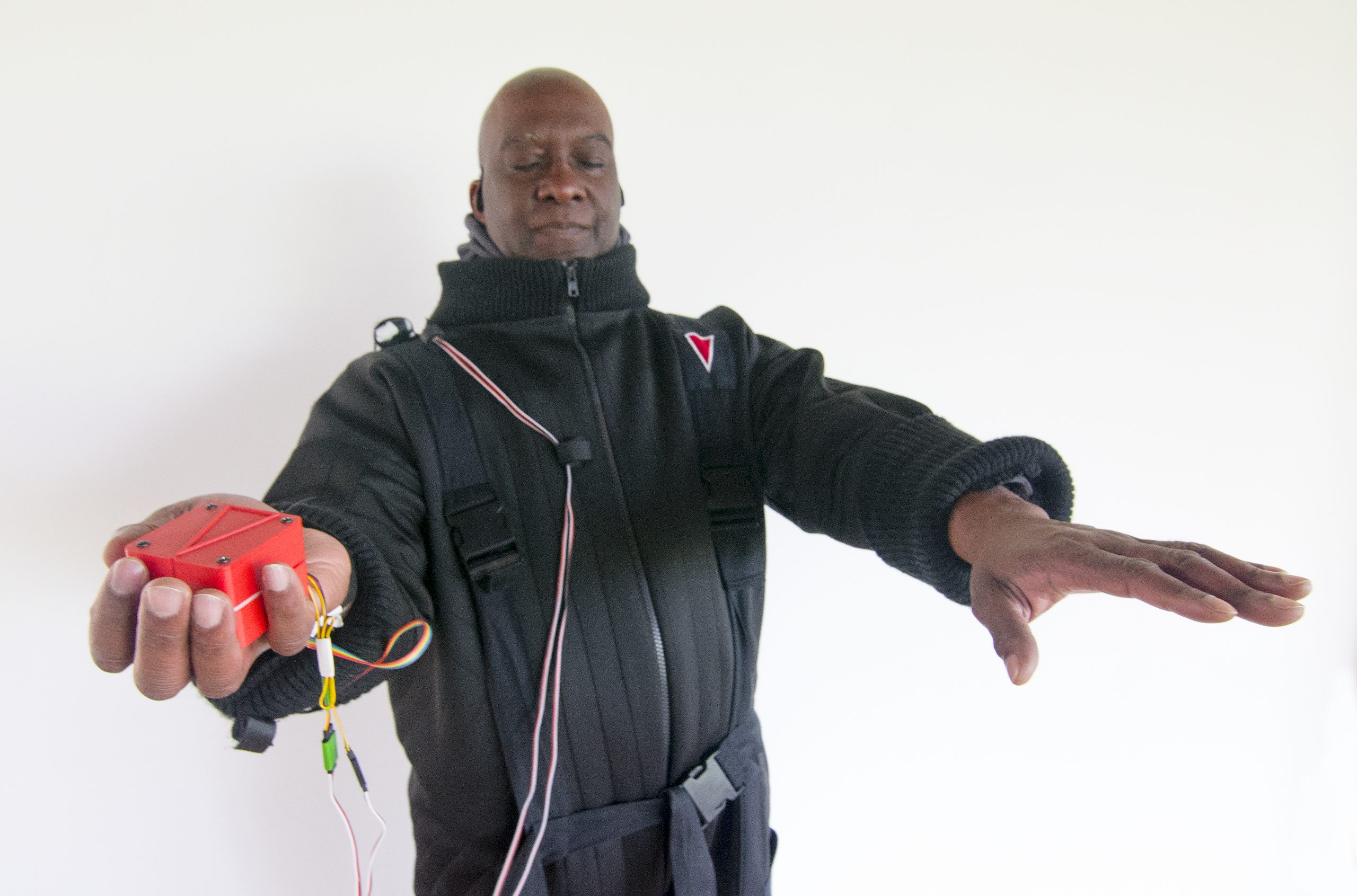 A colour photograph of a tall black man with his eyes closed wearing the black Flatland padded and quilted adventure   jump suit.It has  deep cuffs, a large stand-up collar, and harness like straps and buckles. His arms are raised, with one holding a red Animotus cube with redd and white wires extending out of it and over his chest and shoulder.  The other hand has the palm down reaching out ahead of him.