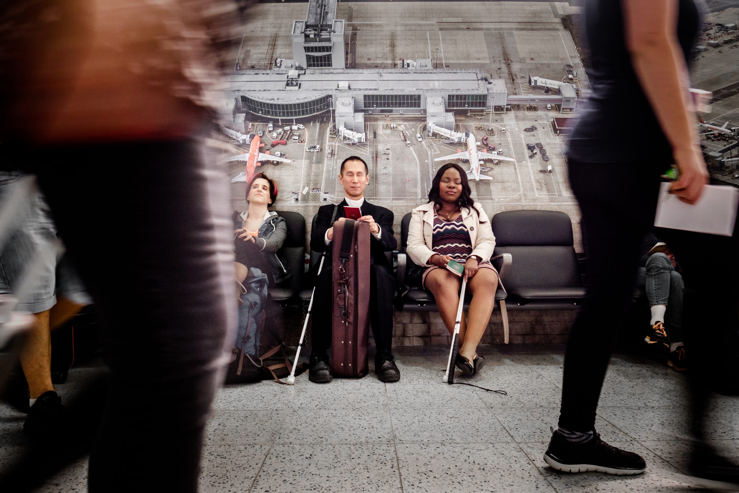 A photograph of two blurred passersby in the foreground, between whom three cast members are in the mid ground in sharp focus. Far left is Amelia, Caucasian, with red tinted dark pixie- cut hair, Takashi   is in the middle, a Southeast Asian man with black cropped hair, clean shaven, wearing black, with a purple viola case between his legs. Victoria, a mid brown skinned   Nigerian woman, with shoulder length wavey black hair, wears a cream jacket and a zig zag patterned dress.Takashi and Victoria hold passports and have resting white canes. all have their eyes closed. Above them is a large 6ft landscape photograph of two jets on the tarmac connected to respective gangways.