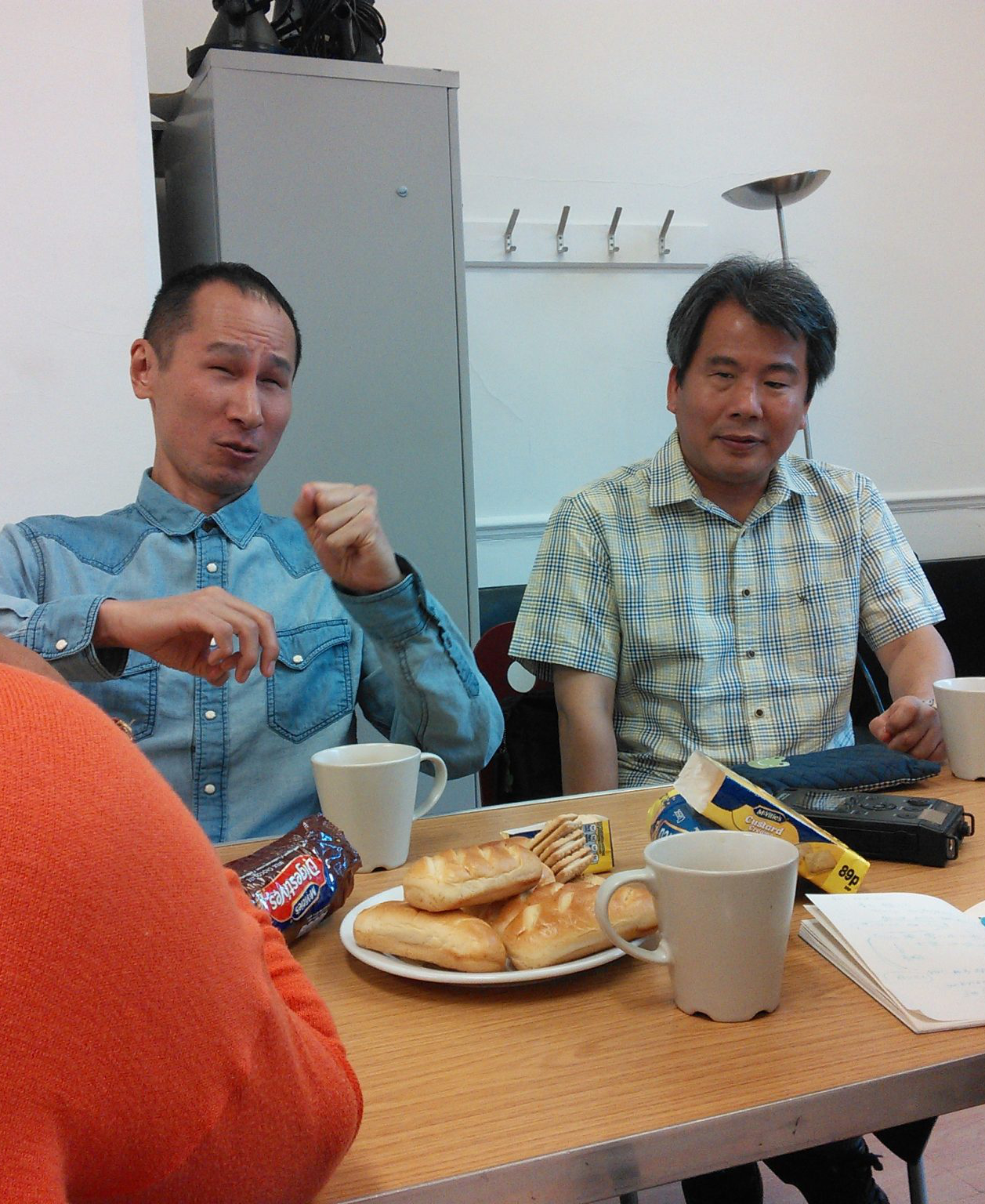 A Photograph of Takashi on the left and Professor Hirose on the right. Both South East Asian middle aged men are sitting at a table on which there are mugs, plates with biscuits and buns, and a recording device. Takashi is in a denim blue shirt facing to camera and gesturing with one hand in a fist and the other curled, while Prof Hirose, also facing front, listens to him