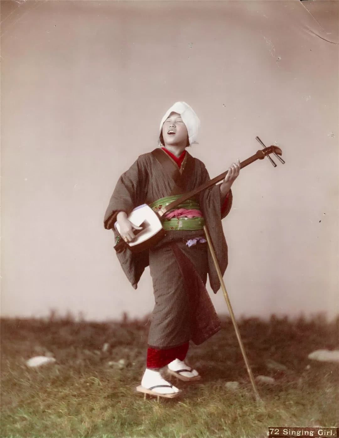 A black and white photograph which has been colour tinted by hand - It is of a Goze performer standing Against a course grass and stoney landscape.  In three quarters profile, she wears an ankle length red dress, over which is a thick outdoor muddy coloured coat. She wears a lime green cloth tied around the waist of the coat with a further pink belt on top.  leaning against her waist is a wooden staff angled out to rest on the ground in front of her, and she wears white socks and simple brown strapped wooden Japanese sandals. Carrying a 3 stringed shamisen instrument, she holds it like a guitar, and her head, which has a white head-wrap, is thrown back as she sings.   