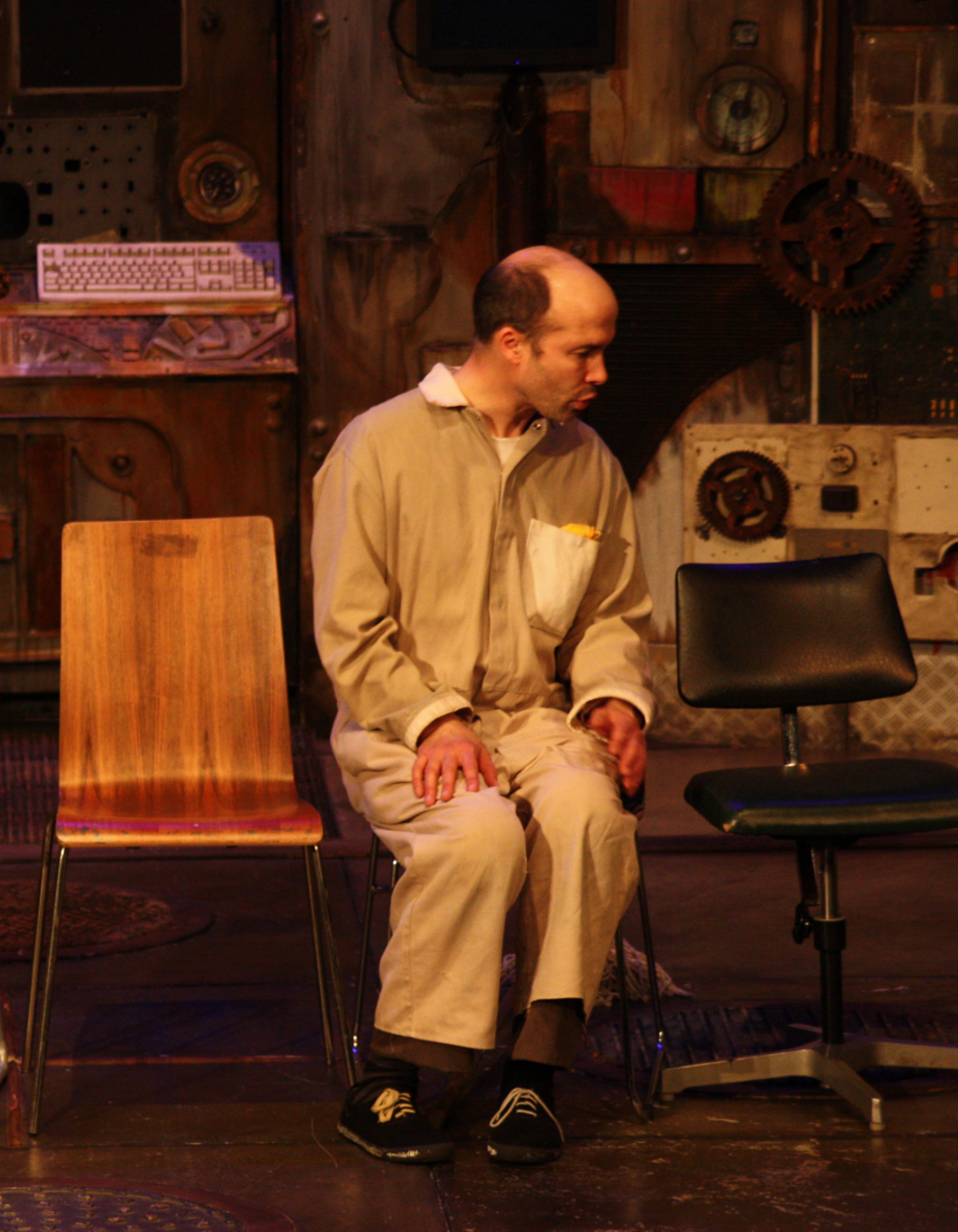 A photograph of Tim Gebbles as the Old Man, sits in overalls on one of three chairs, two of which are empty.  His head is turned to the right and he is speaking to an invisible character next to him.