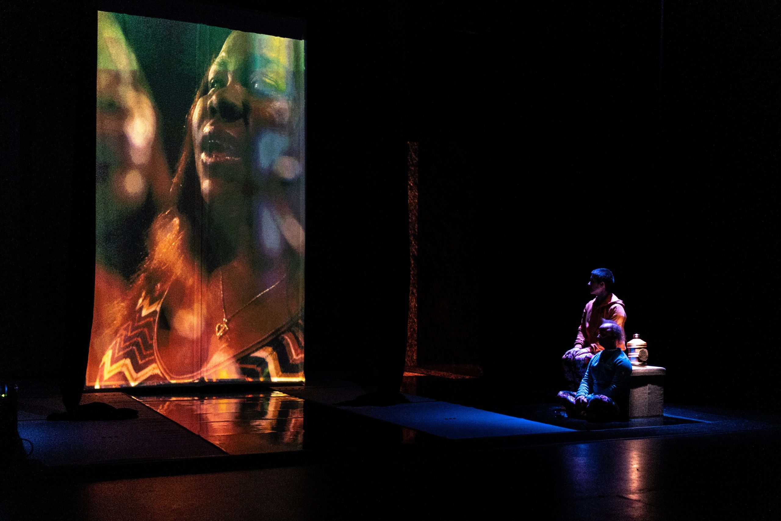 A photograph - Against a dark background two white performers, Amelia and Sarah, sit partially lit stage right. One is in pink and the other in blue, and they both focus on a 14foot portrait screen, on which Victoria, a black woman, sings in profile.  On the bottom half of the screen there is a golden glow from lighting, and a greenish glow at the top.  This effect is reflected in the black lacquered floor of the set.