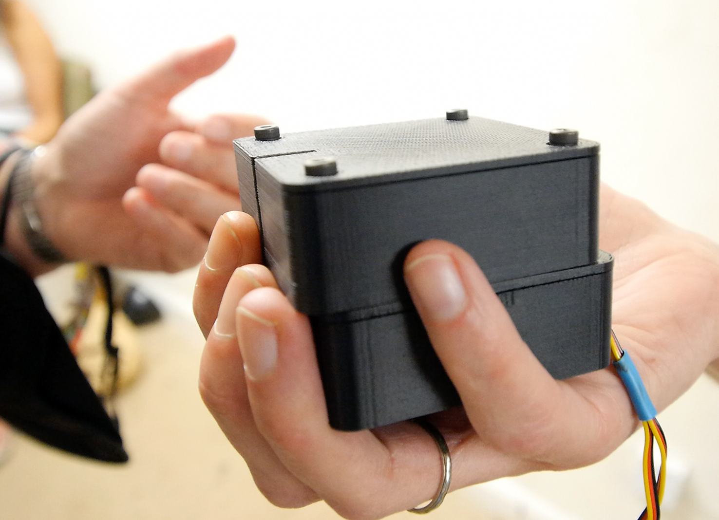 A close up of a white male hand side on holding a black Animotus cube which sits in the palm. The fingers are lightly curled up around the cube and the top half of the cube is slightly swivelled to the right. 
Protactile Description – The Animotus cube has a slightly grainy textured feel to the surface and is as heavy as a Rubrik’s cube. It vibrates mildly when activated and, like the Lotus, makes a quick switching sound when swivelling from left to right or extending straight ahead.