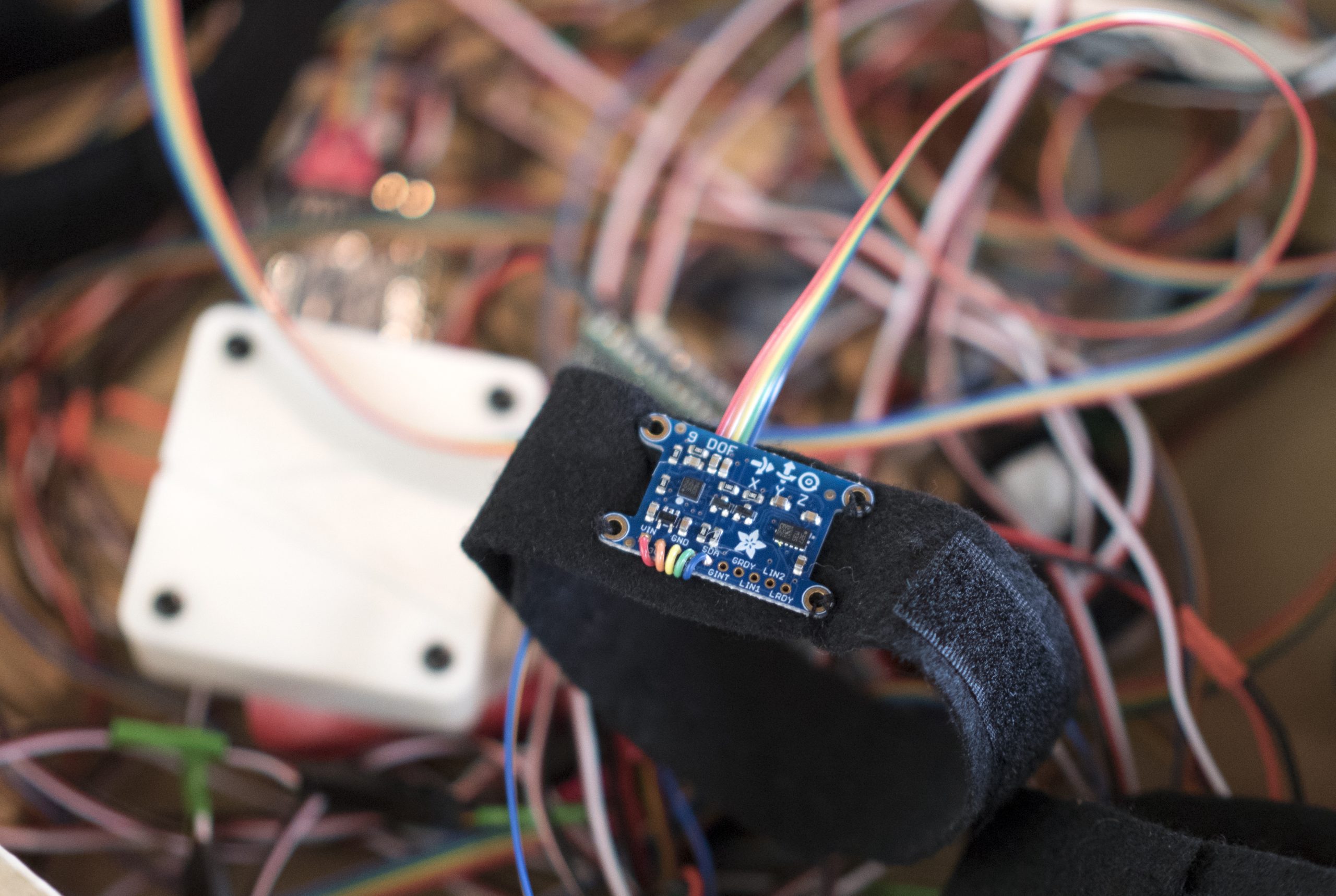 A photograph of a Black Velcro wristband attached to a circuit board with red, orange, green and blue wires leading into a cable that is part of a blurred tangled heap of cables, that the magnetometer rests on