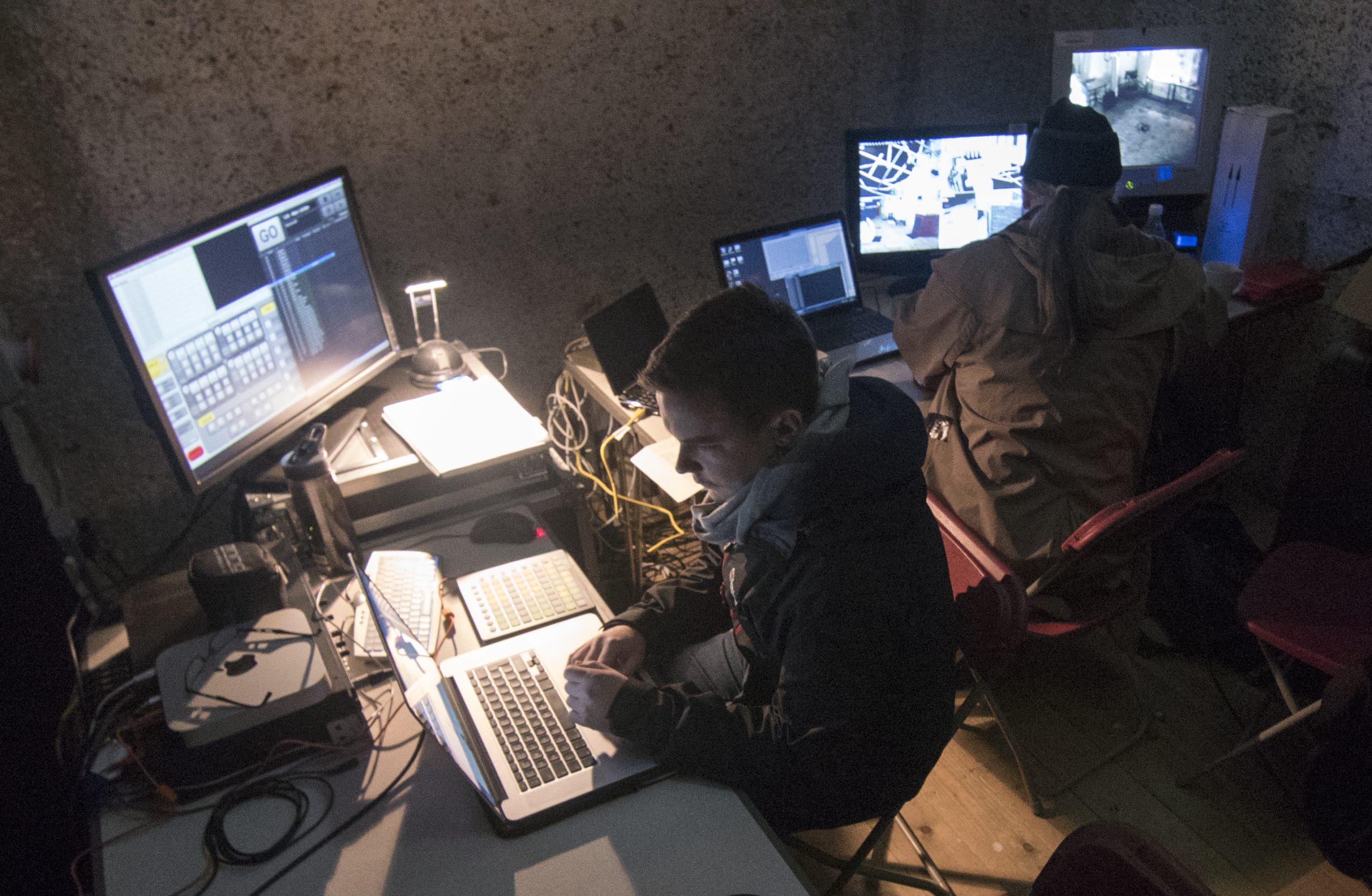 A photograph of a younger and older white male, seated in front of laptops and 3 monitors, with additional keyboards and screens in front of them. The monitors show black and white subdivisions of an interior space and other shows columns of lots of coding data. 