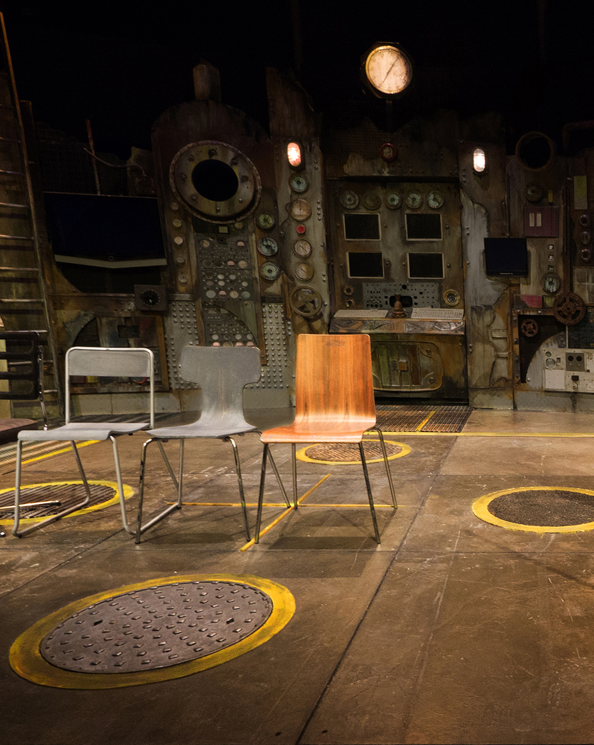 A photograph of the floor of the set lit with the dimmed walls of the backdrop in the background.  In the centre are three different utilitarian chairs in a row facing front. Under the chairs is a vivid yellow cross, and at the end of each yellow strip, North, South, East and West, is a round circular metal looking cover, each with a different texture surface.  One has bumps, two have a ribbed surface, and the fourth has a granular texture.  At the back, in the centre of the walls of the power station is a central panel and on the floor in front of this is a rectangular ribbed panel with a yellow strip running in front of it.  On the extreme left-hand side of the floor area is a narrow carpet strip running from downstage to meet the power station wall at the back with yellow strips at regular intervals.