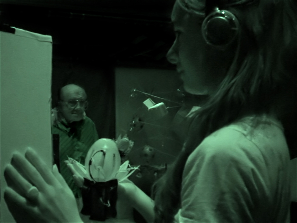 A monochrome image taken with an infra red camera, so a dull muted olive-green filter covers everything. On the right, in profile, is a young woman, wearing headphones, holding a Lotus in her right hand with her left hand touching one surface of a large cube hanging in front of her. In the background another traveler holding a Lotus is in motion