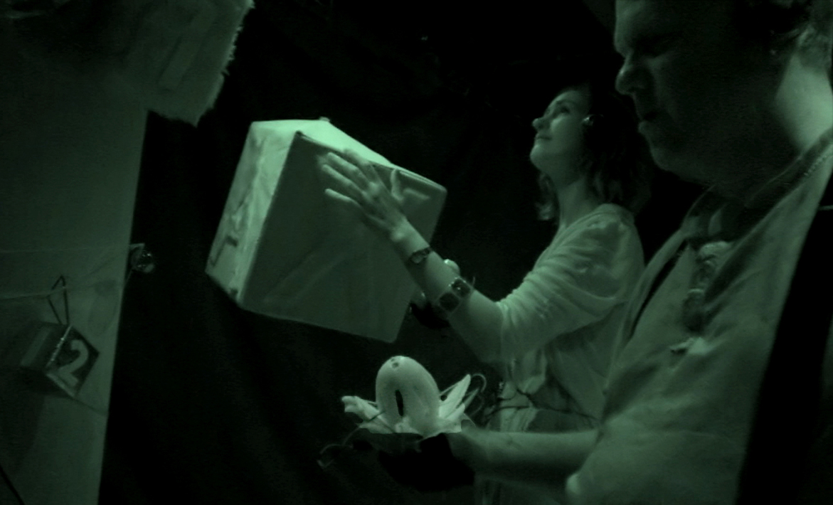 An infra red photograph of a man in the right foreground facing left holding out the lotus while a woman in the background stands raising her hand up to feel a cube hanging in front of her.