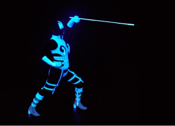 A figure stands in black light with glowing white aspects of costume picking out their fencing pose – side on to the camera, legs wide apart, knees bent, right arm on right hip and left arm above their head pointing forwards holding a glowing white cane thrust forward like a rapier.