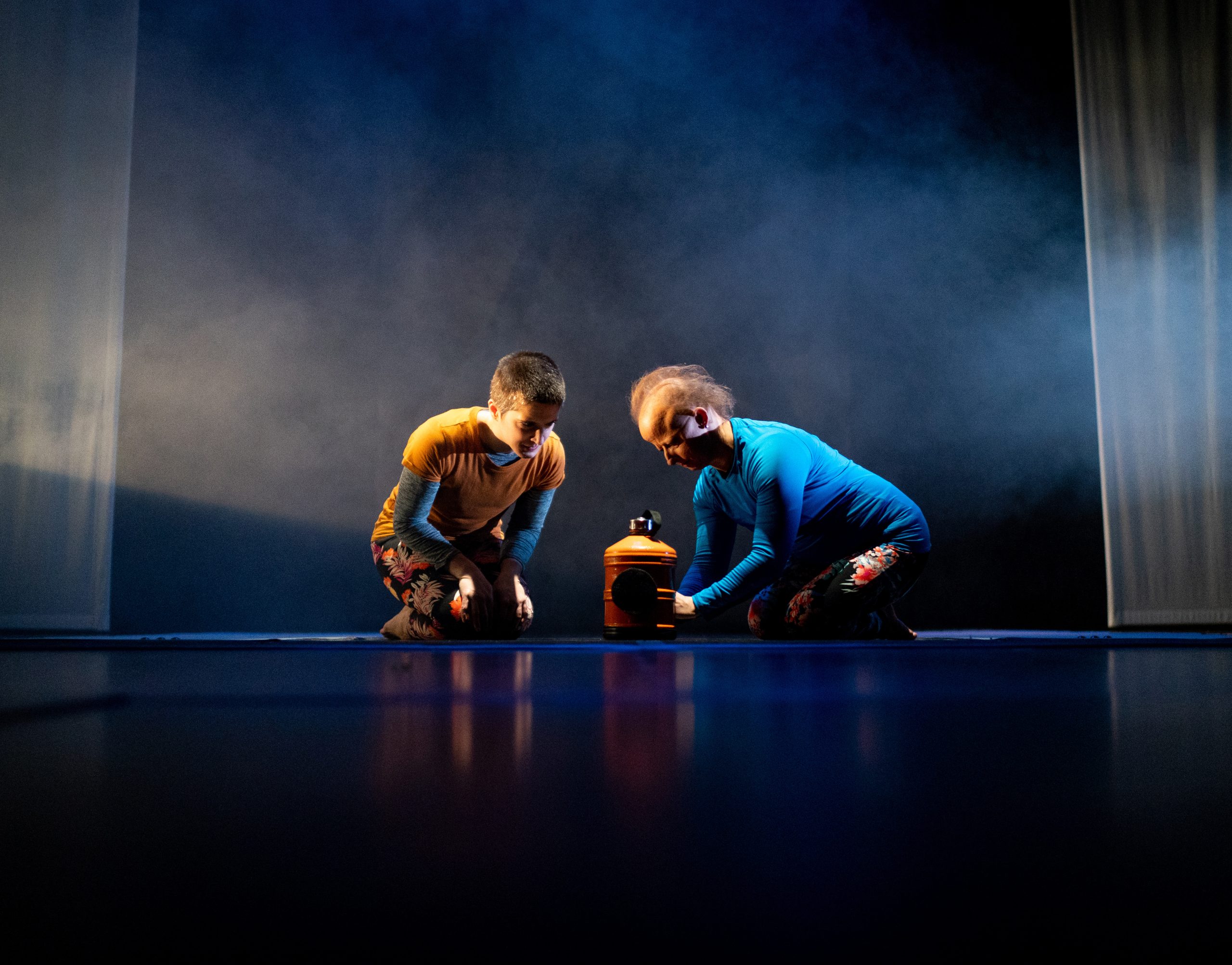 A photograph - Against a misty blue lit background, Amelia on the left in an orange top and Sarah on the right, in a blue top, both kneel on their haunches, leaning forward and focus intently on a large orange water bottle between them. Sarah is extending her arms out to it.   