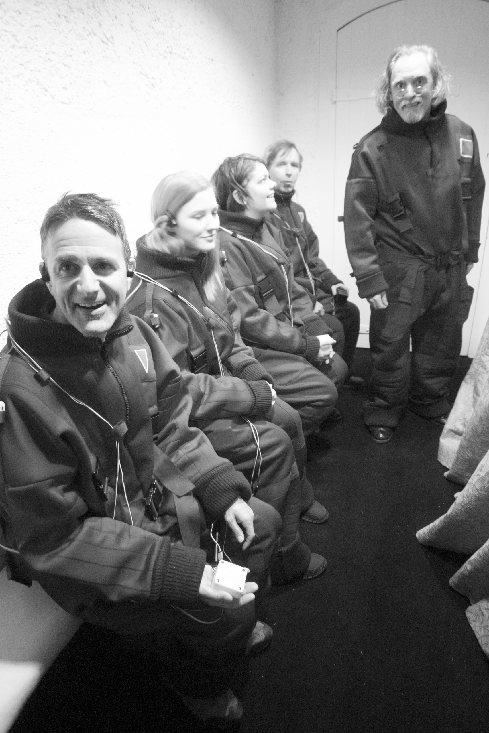 A photograph of four people sitting on a bench against a wall, all wearing black padded Flatland jump suits. Some are holding the Animotus in their hand and all appear animated, while David Fielder as Elder Square stands next to them in a Flatland suit, looking out to camera