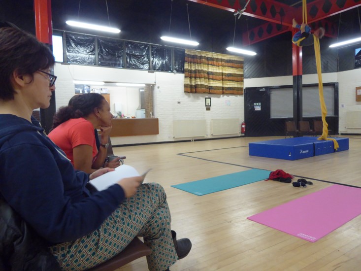A photograph of a vaulted rehearsal room   with a red supporting beam structure and pink and blue exercise mats laid out on a wooden floor. In the foreground Kumiko   in profile with dark hair tied up, sits back in an upright chair wearing a dark blue patterned top with her two hands holding an open notebook on her lap. Maria to her side and further back, in a   reddish short sleeved t shirt, sits in a chair leaning forward. Both are focused intently on Sarah hanging in a yellow silk at the other end of the room, in a fetal position with her legs apart. 