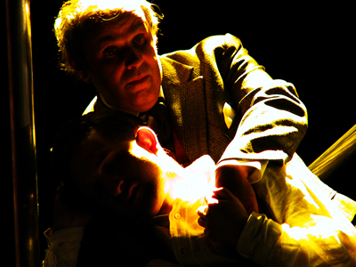 Against a dark background, lit by an orange glowing light, older Jacques cradles younger Jacques in the scaffold.