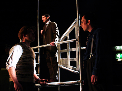 In the foreground Francois, in sleeveless jumper, shirt and flat cap, faces Jean in tie and braces, with professor standing up high on scaffold behind them.