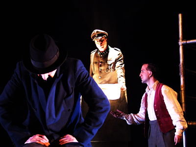 A new recruit in foreground sitting in a chair facing out, lit by a bluey light from below, dressed in trench coat with up turned collar, head bent, wearing a trilby hat. The Major is behind on a podium, lit by an orange light from below and younger Jacques caught in the podium light on the right, gestures towards Denis with a hand.