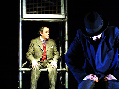 The new recruit in foreground in same position as in shot 19, lit by a blue grey light, now with older Jacques sitting behind him on the scaffold, which is picked out in bright orange light, against a dark background. 