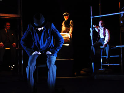 Older and younger Jacques sitting left and right on Scaffold set, with the Major on a podium lit from below by a yellow light, and new recruit in fore ground, sitting in chair, facing forward, head Lowered, bathed in blue and black shadows.