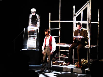 Major, back left, reads long report trailing over front of his Podium, while teacher sits on brightly lit scaffolding set to the right in a brown suit, reading a document and younger Jacques in the left of the fore ground, standing with head bent