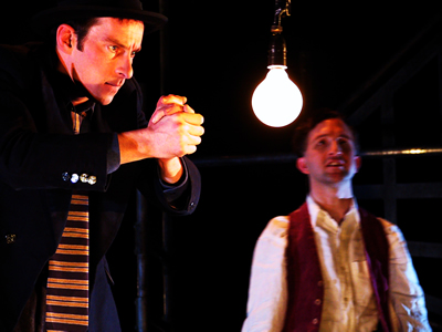 Denies in brown trench coat and trilby hat standing on left, hunching towards and warming hands over large light bulb suspended between him and younger Jacques, who stands on right, facing out.