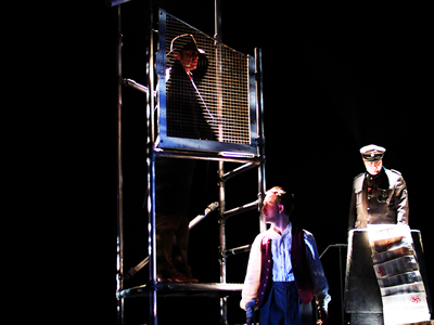 Distant shot of image in 29 - Full length of Philippe on scaffold, face behind wire mesh, with younger Jacques standing against scaffold below, facing out, and Major on his podium lit up from below in orange light.