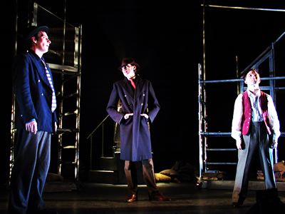In front of both scaffolded towers, in foreground on left, Denis profiled in blue jacket and trilby, looking towards younger Jacques, standing on right facing out with legs apart and head slightly back. Between them, the tall figure of Philippe stands with hands in pocket of brown over coat, looking at Denis.