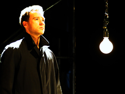 Waist up shot of Eliot on left facing out in a buttoned up overcoat, collar turned up and trilby hat, a yellowish light revealing his face. On the right is the large light bulb suspended in the air.